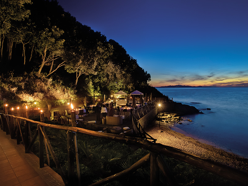 Outdoor seaside restaurant illuminated at dusk with a view of the ocean and surrounding forest.