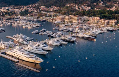 Aerial view of luxury yachts docked at Porto Montenegro marina with a coastal town and forested hills in the background.