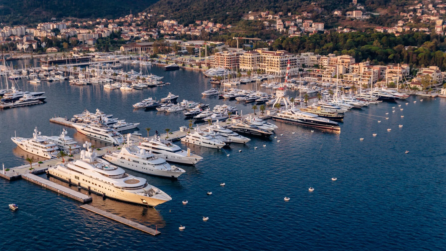 Aerial view of luxury yachts docked at Porto Montenegro marina with a coastal town and forested hills in the background.