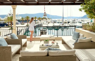 Eine Frau steht auf einer Terrasse mit Blick auf einen Yachthafen voller Yachten, umgeben von Palmen.