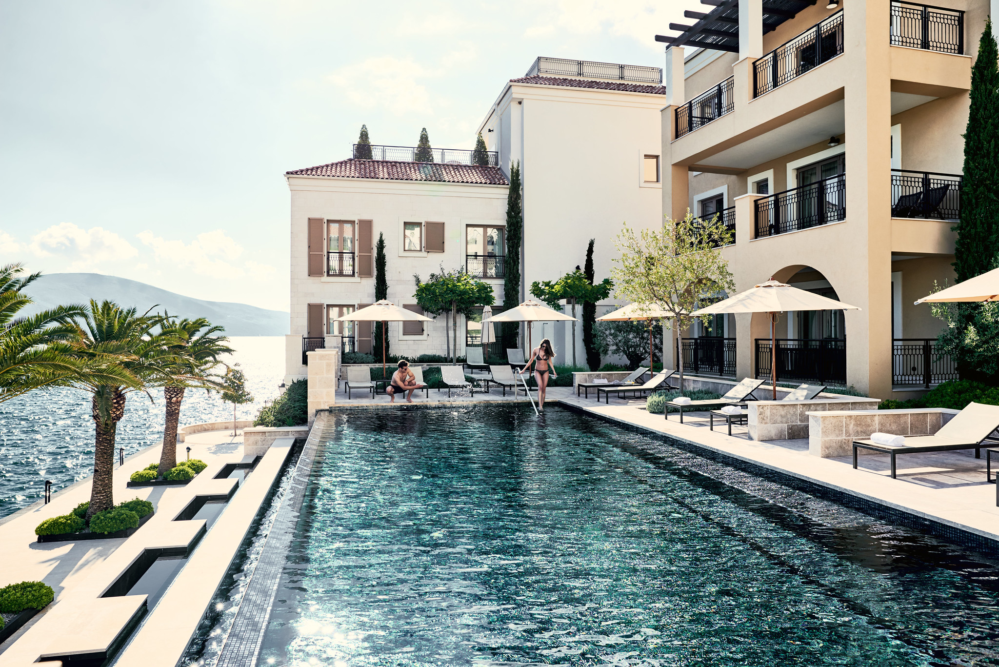 Luxury seaside resort with a pool, sun loungers, and a woman entering the water, set against a backdrop of elegant buildings and palm trees.