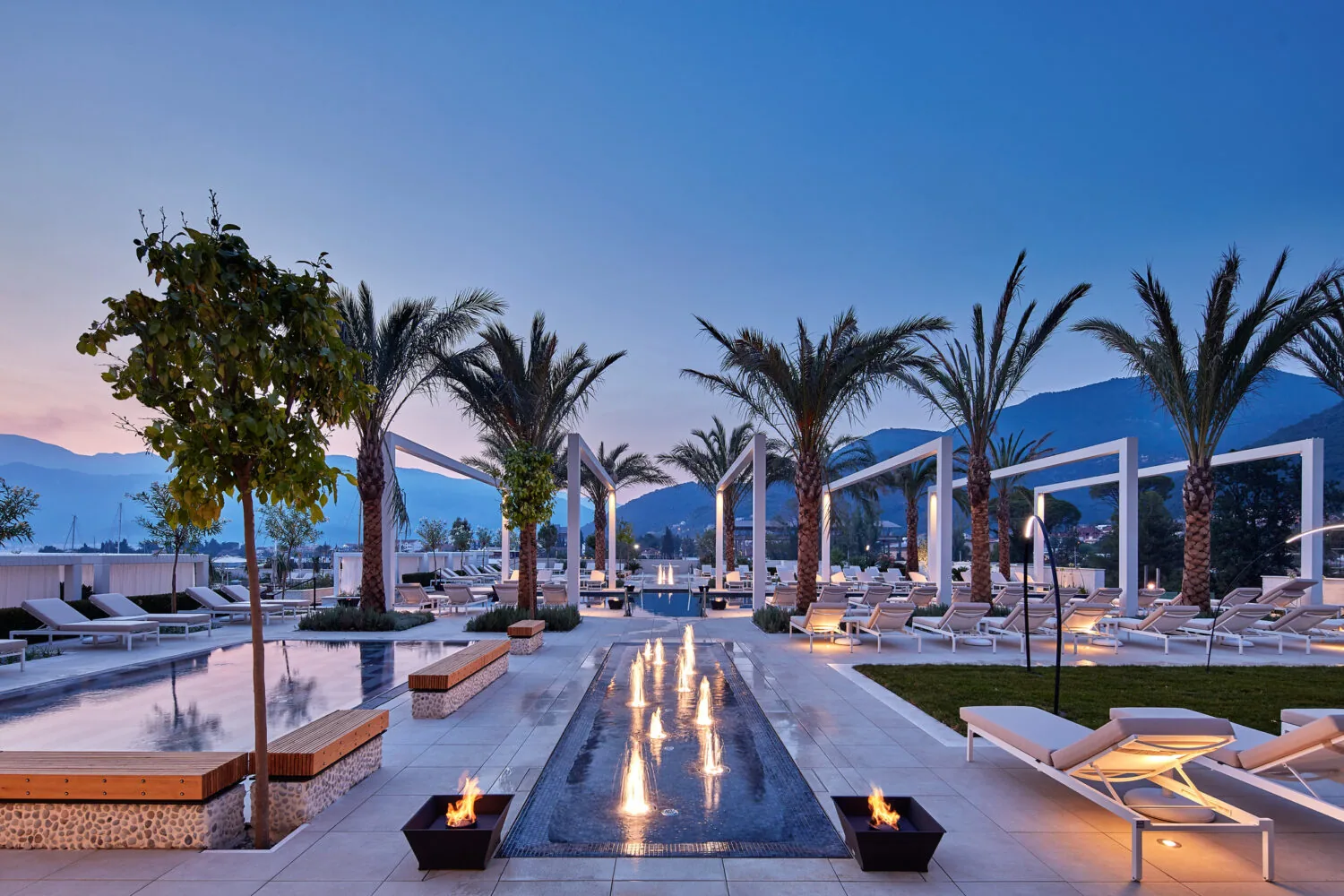 Modern luxury resort pool area at sunset with palm trees, fire features, and lounge chairs.