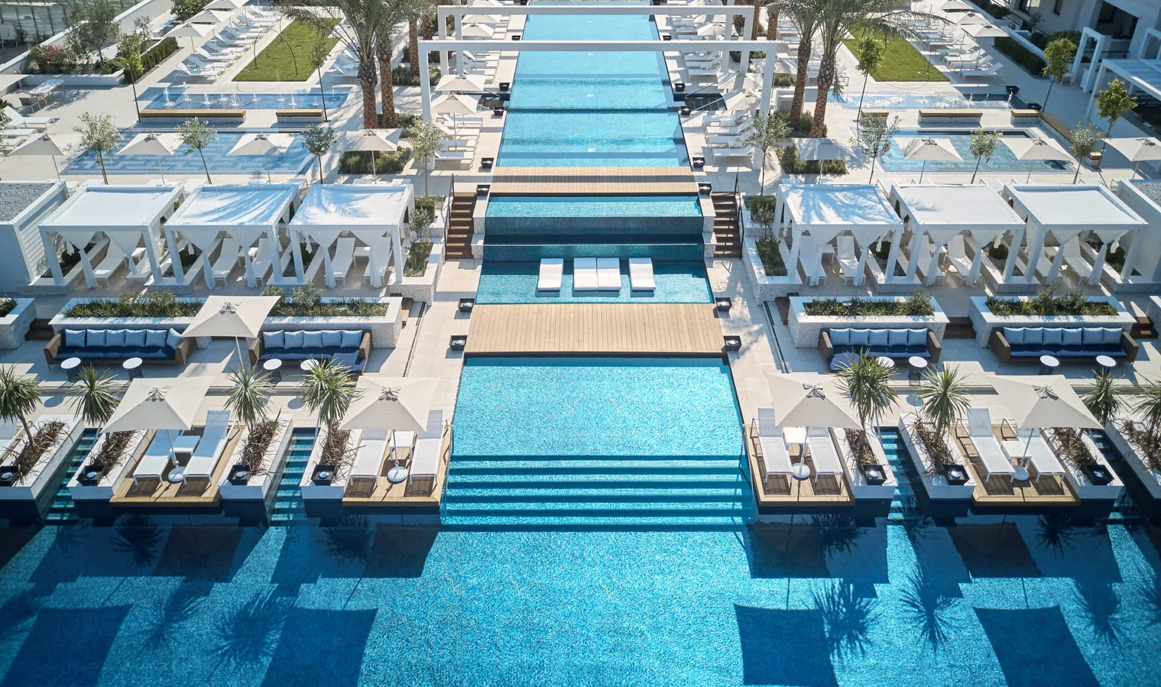 Luxurious resort pool area with multiple tiered infinity pools, cabanas, lounge chairs, and palm trees.