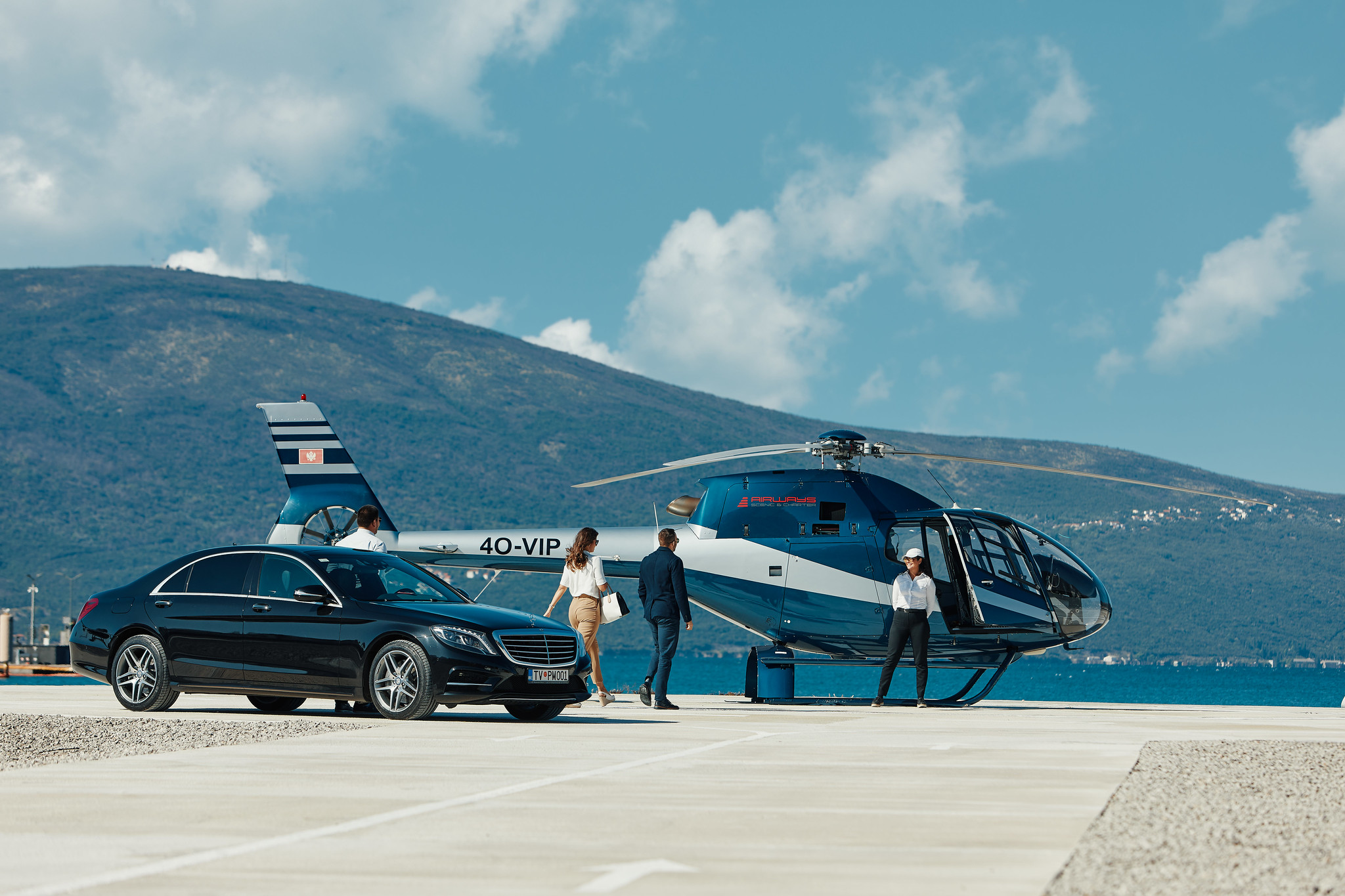 Luxury travelers walk from a black Mercedes-Benz to a VIP helicopter on a sunny helipad near the coast.