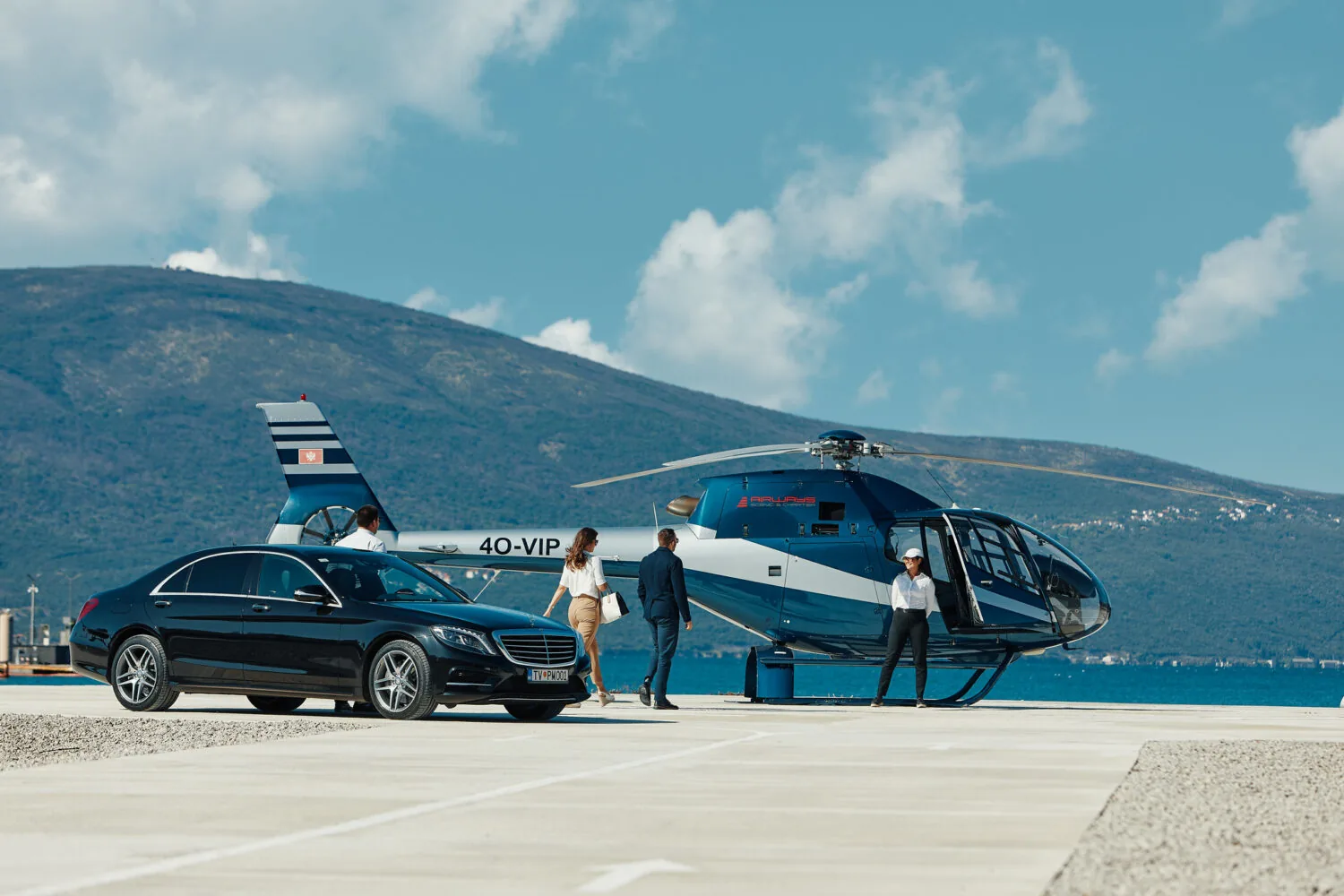 Luxury travelers walk from a black Mercedes-Benz to a VIP helicopter on a sunny helipad near the coast.