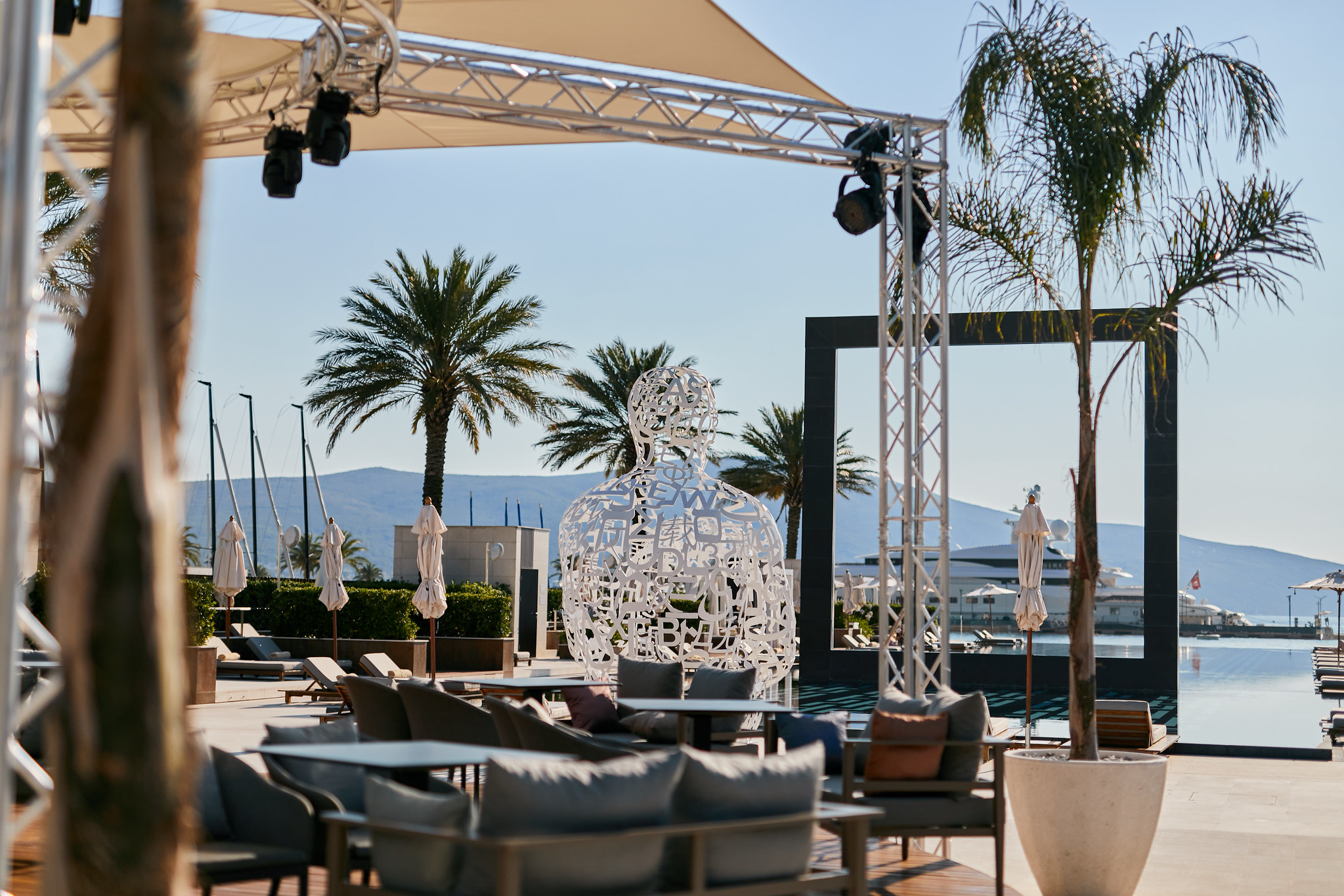 Outdoor lounge area with modern seating, palm trees, a white alphabet sculpture, and views of a marina and mountains in the background.