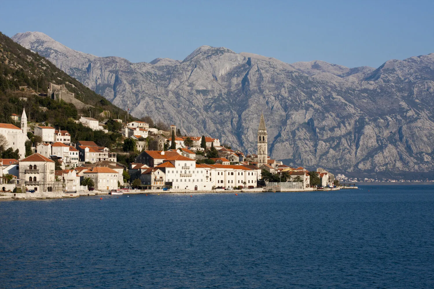 Coastal town of Perast in Montenegro with historic buildings, a church tower, and a mountainous backdrop by the Bay of Kotor.