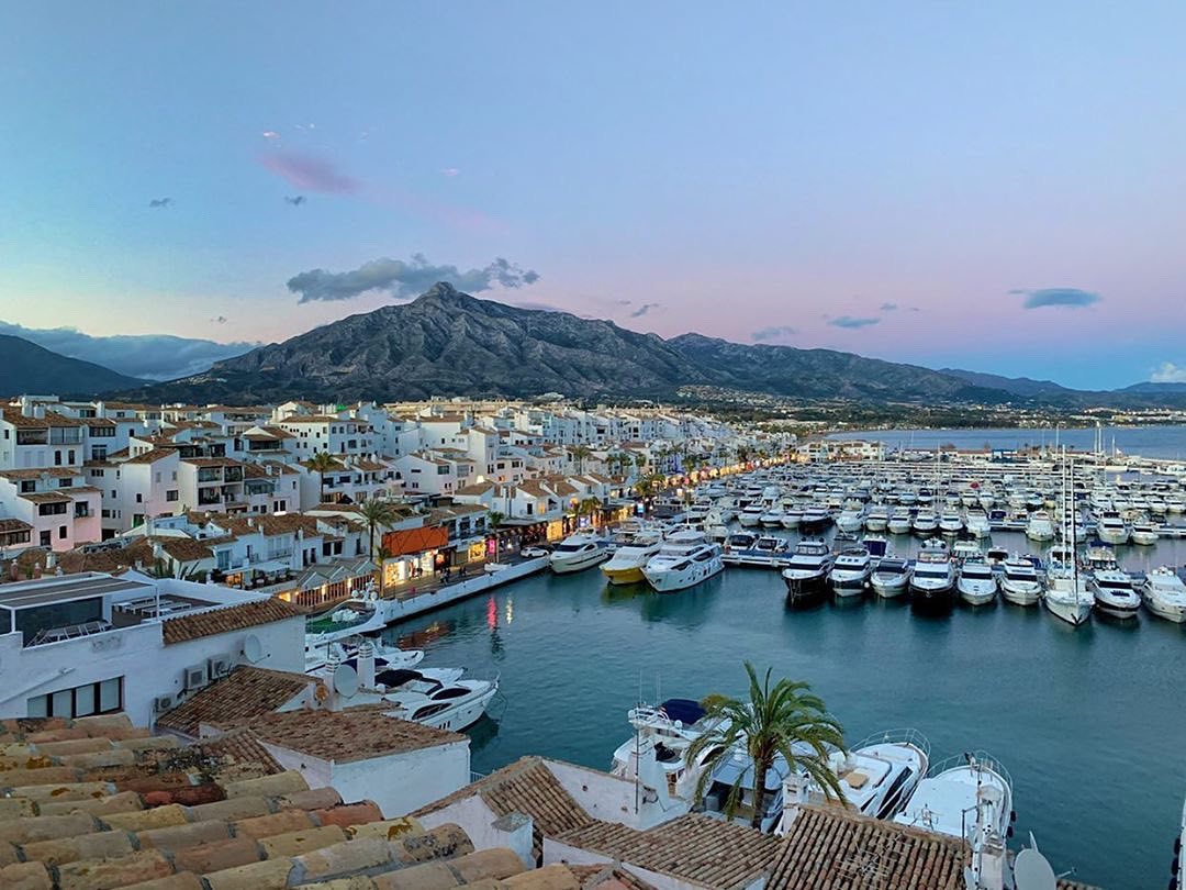 View of Puerto Banús marina at sunset with numerous yachts and white buildings set against a mountain backdrop.