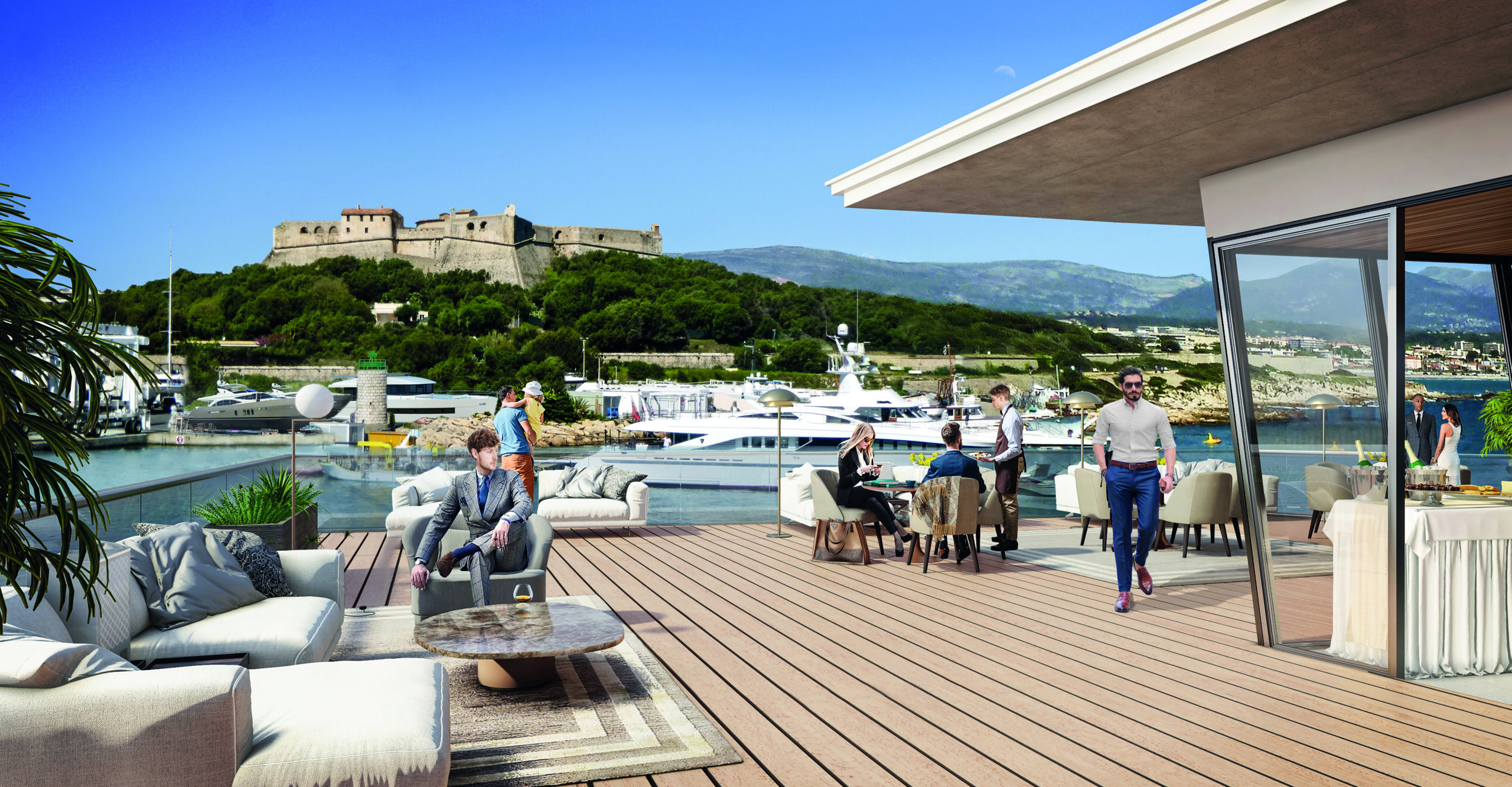 People relaxing and socializing on the IYCA terrace with yachts in the harbor and Fort Carré visible in the background.