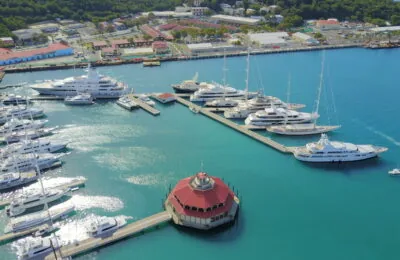 Aerial view of a marina with numerous luxury yachts docked in turquoise water near a coastal town.