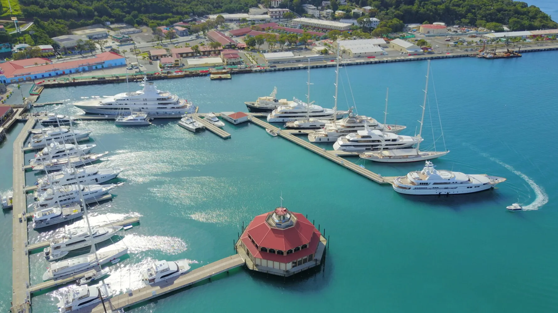 Aerial view of a marina with numerous luxury yachts docked in turquoise water near a coastal town.