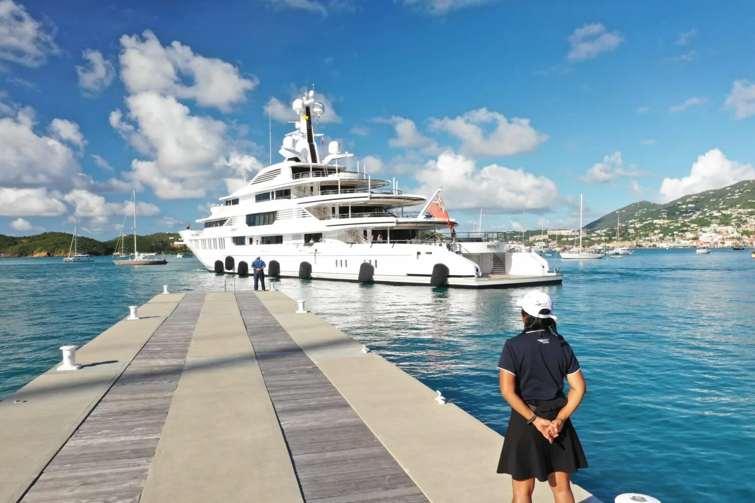 A luxury superyacht is docked at a marina while crew members stand on the pier under a sunny, blue sky.