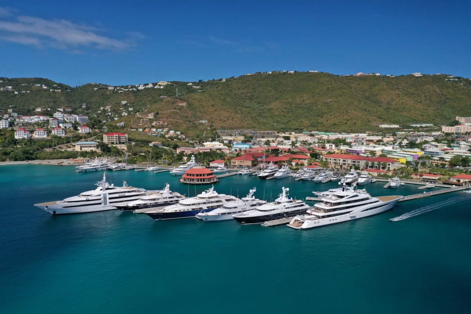 Aerial view of a marina filled with luxury yachts in front of a coastal town surrounded by green hills.