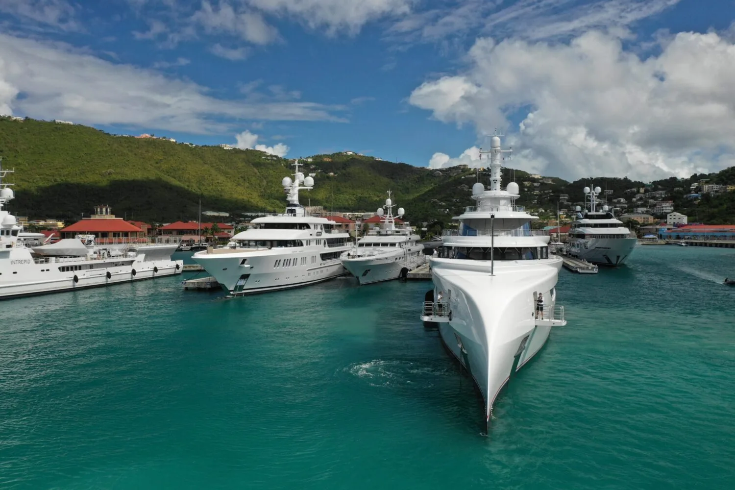 A row of luxury yachts docked in a tropical marina with green hills and colorful buildings in the background.