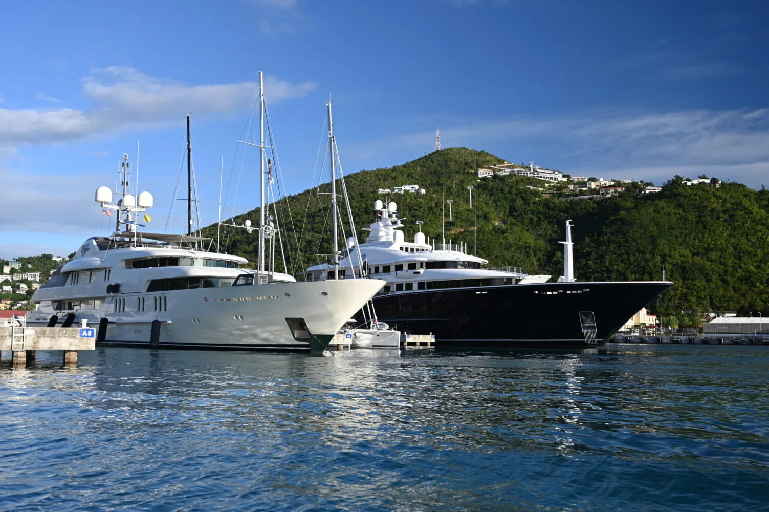 Two large luxury yachts docked at a marina with a green hill and blue sky in the background.