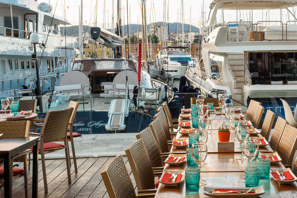 Elegant outdoor dining table set with glassware and red napkins overlooking luxury yachts in a marina.