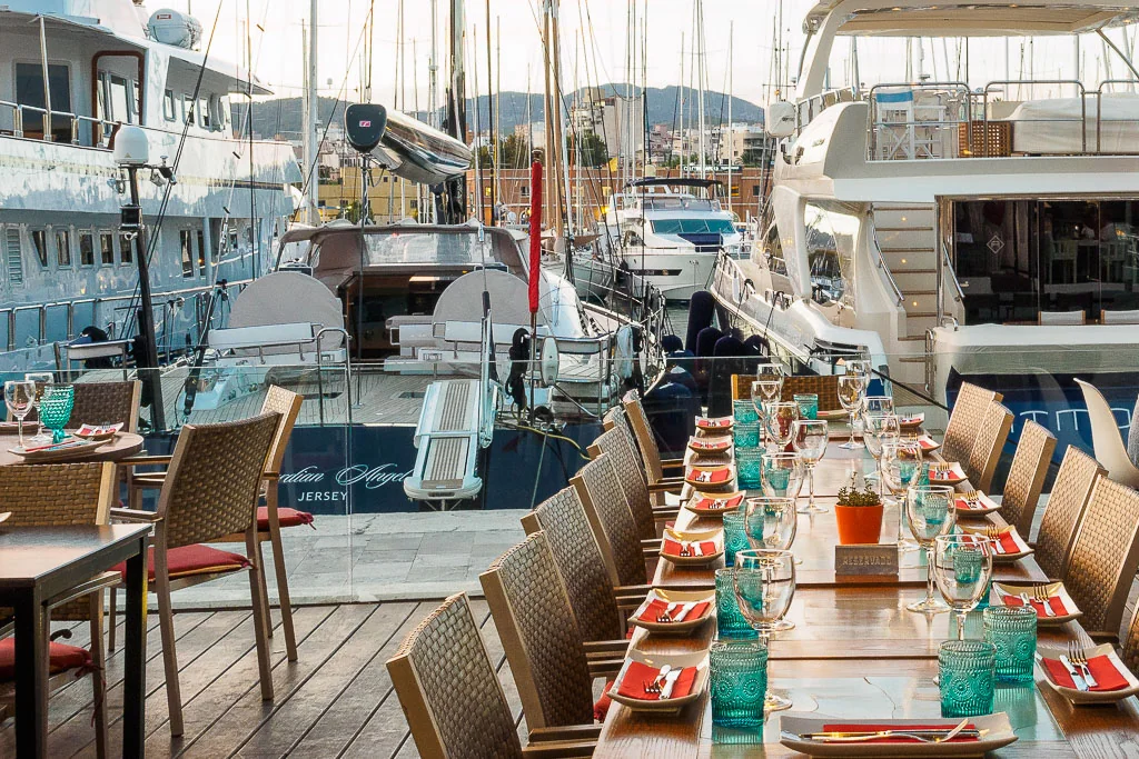 Elegant outdoor dining table set with glassware and red napkins overlooking luxury yachts in a marina.