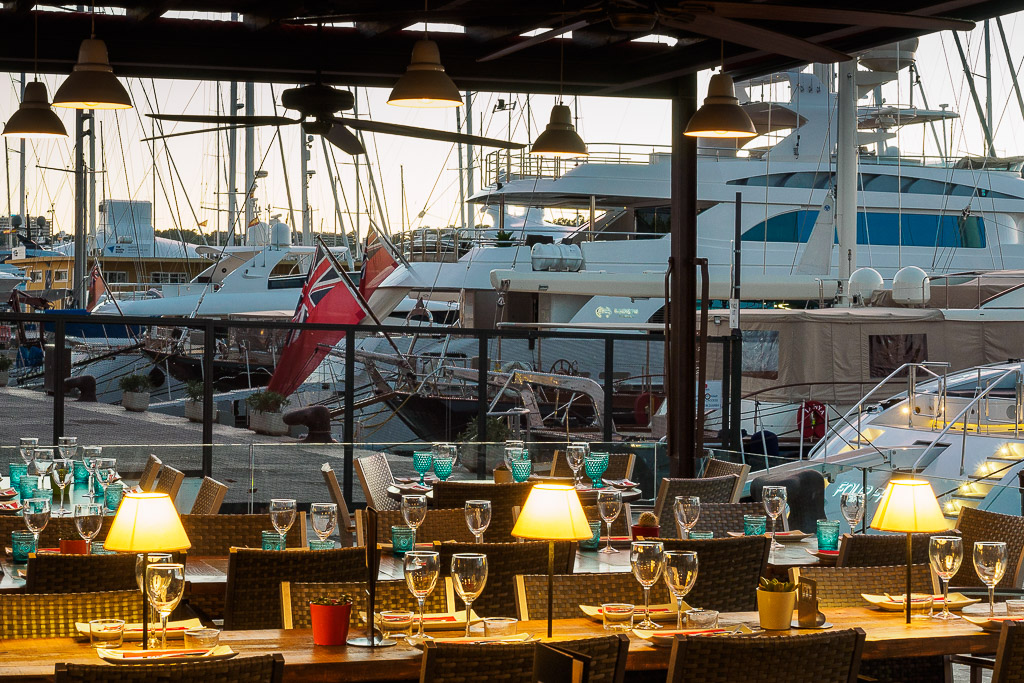 Outdoor restaurant with neatly set tables overlooking a marina filled with luxury yachts at sunset.