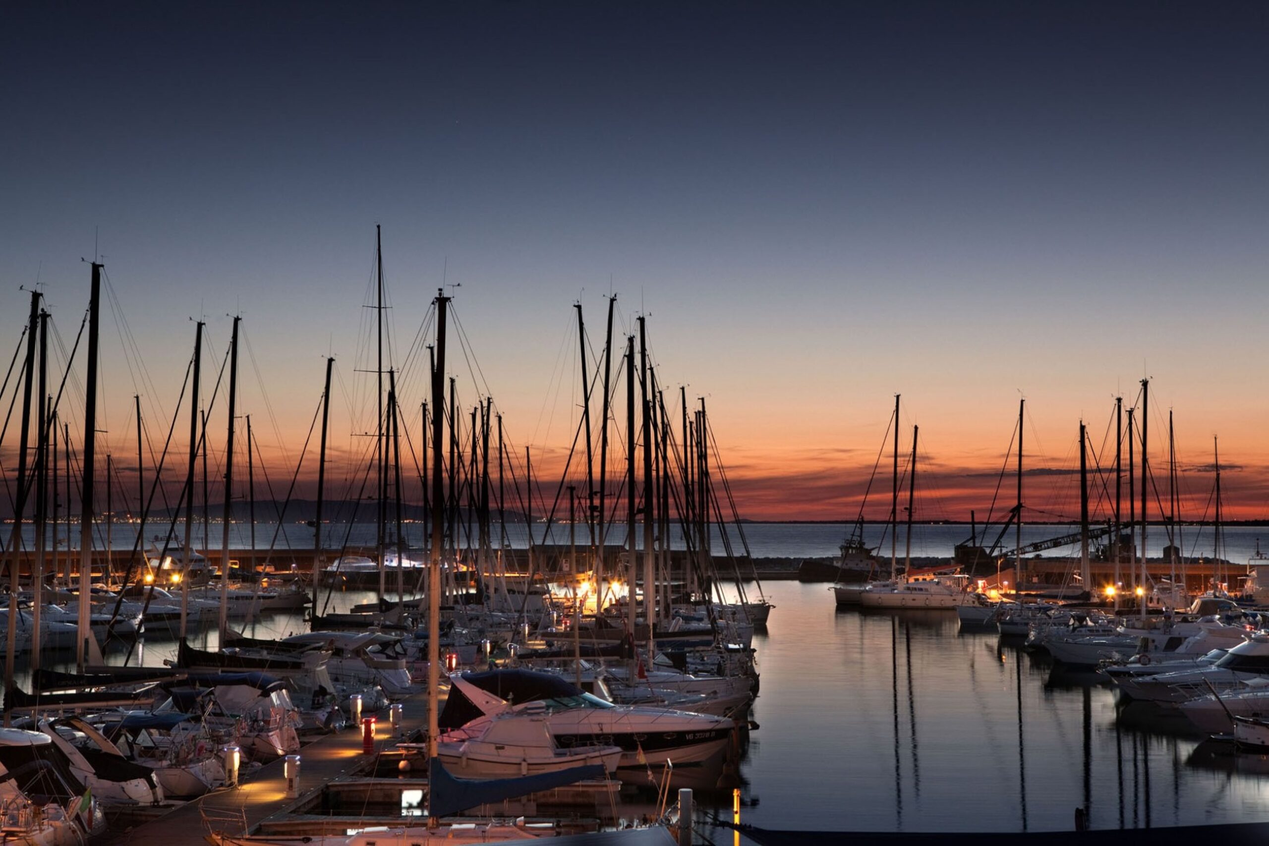 A marina filled with sailboats and yachts at sunset, with calm water and a glowing orange and blue sky.