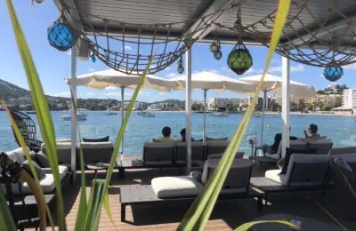 Menschen, die sich auf Liegestühlen unter einer schattigen Terrasse mit Blick auf eine sonnige Meeresbucht mit Booten und entfernten Gebäuden entspannen.
