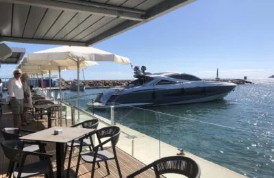 Eine Luxusyacht, die in der Nähe einer Restaurantterrasse mit Sitzgelegenheiten im Freien und Sonnenschirmen angedockt ist.