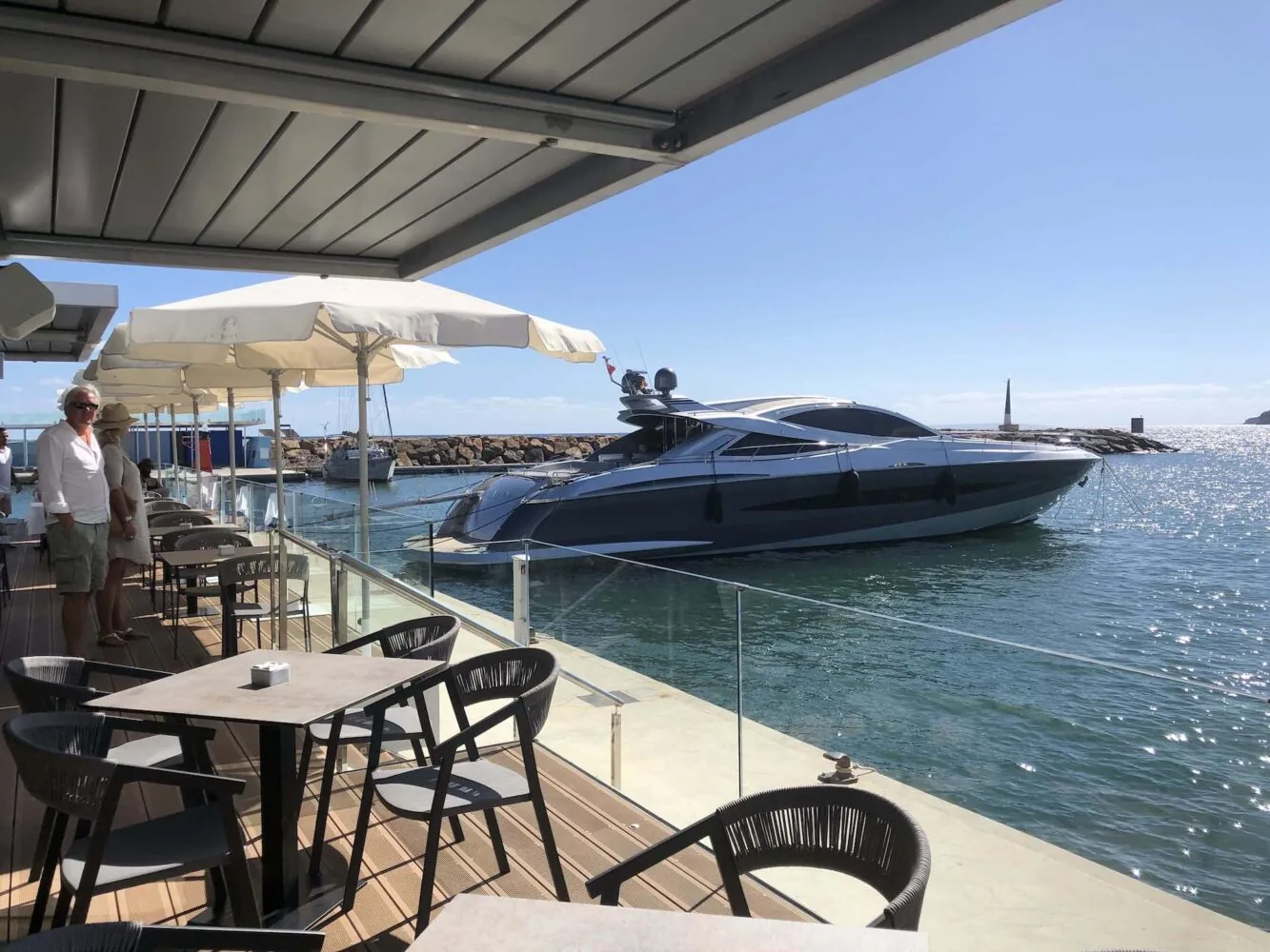Eine Luxusyacht, die in der Nähe einer Restaurantterrasse mit Sitzgelegenheiten im Freien und Sonnenschirmen angedockt ist.