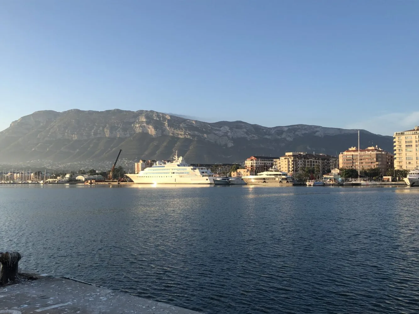 A coastal city marina with yachts docked in front of mountains and apartment buildings at sunset.