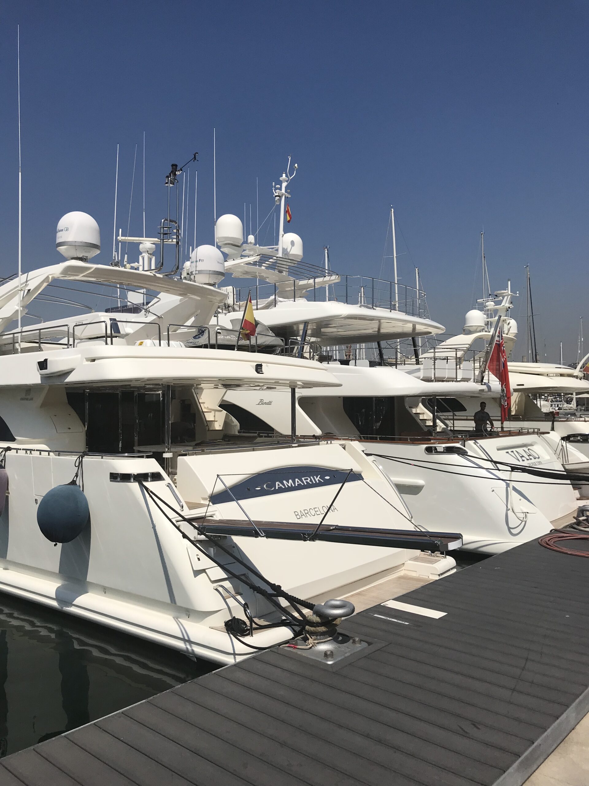 Luxury yachts moored at a marina under a clear blue sky, including one named "Camarik" from Barcelona.
