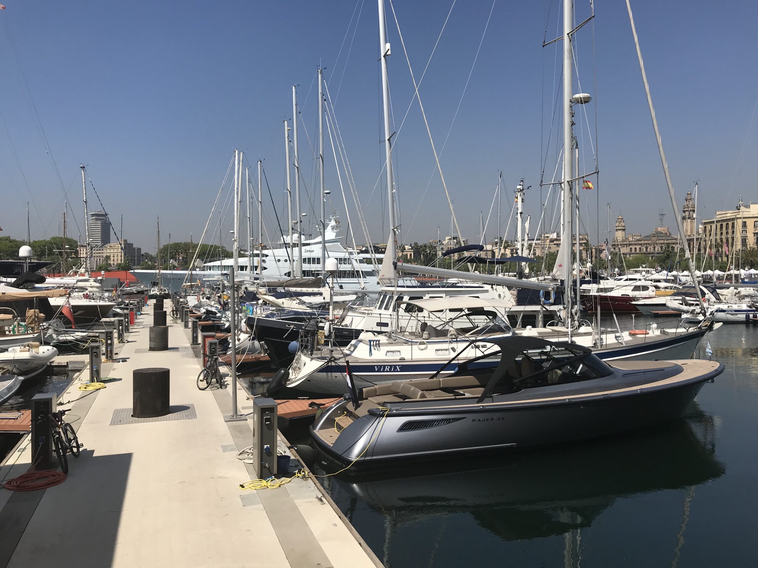 Several yachts and sailboats docked at a sunny marina with buildings visible in the background.