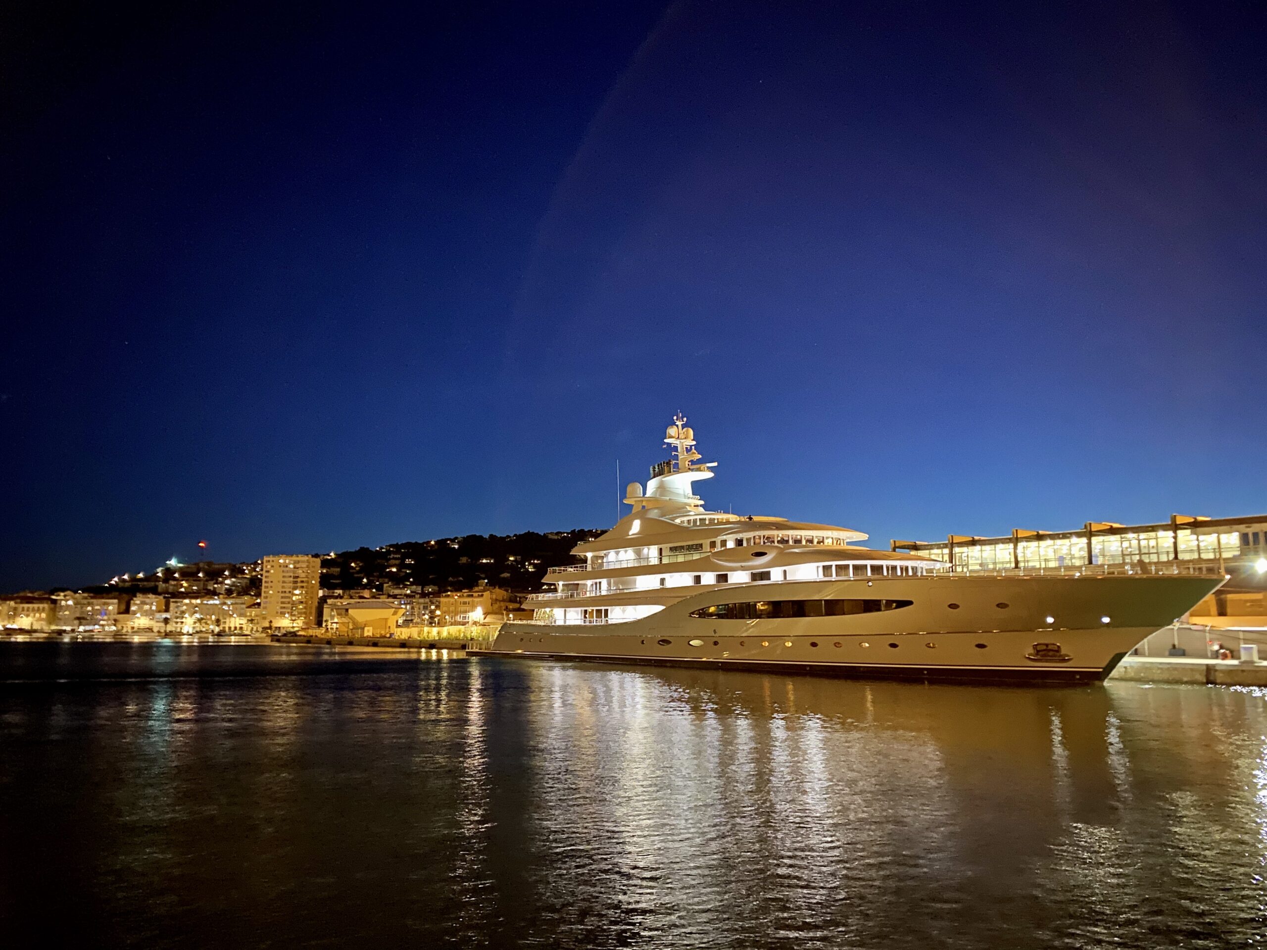 Luxury yacht docked at a brightly lit harbor at night with a hillside town in the background.