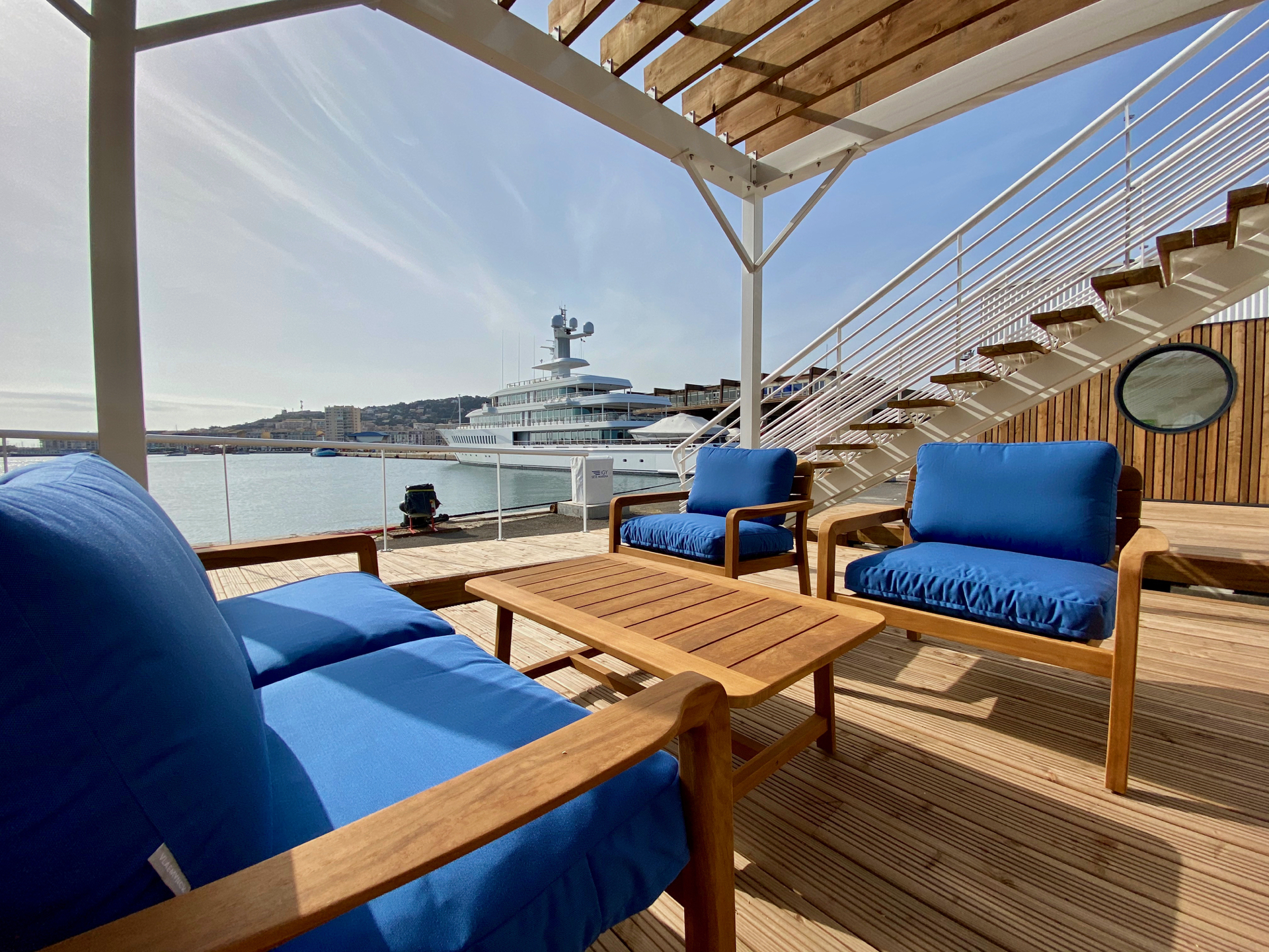 Wooden deck with blue cushioned lounge chairs overlooking a luxury yacht in the harbor.