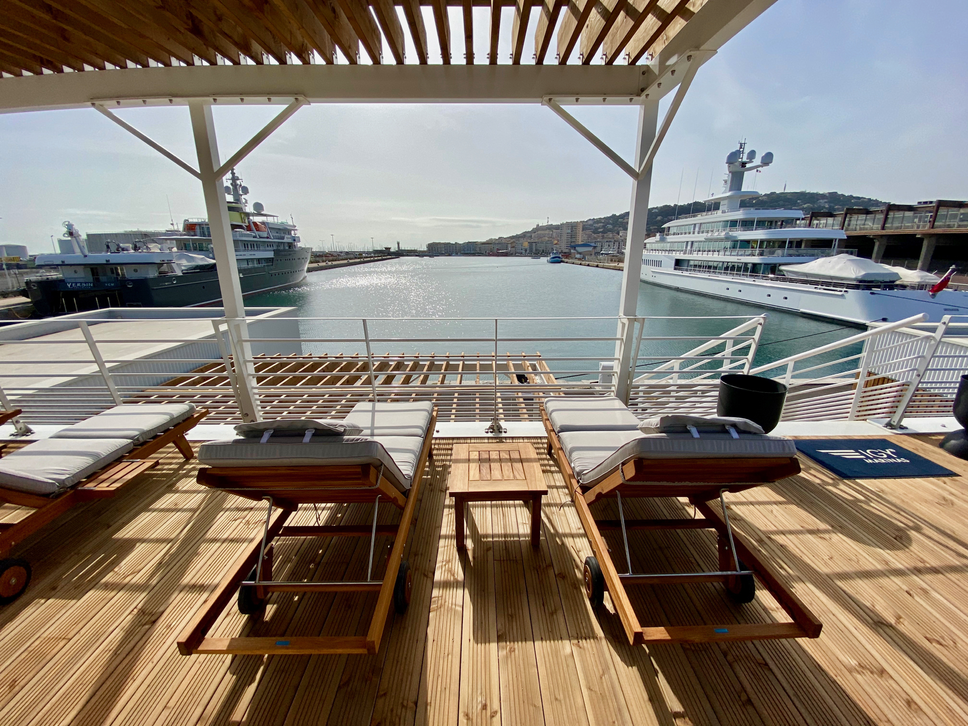 Two sun loungers under a wooden pergola overlook luxury yachts docked in a marina.