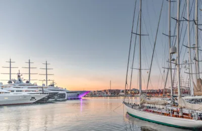 Luxury yachts and sailboats docked in a marina at sunset with city buildings in the background.