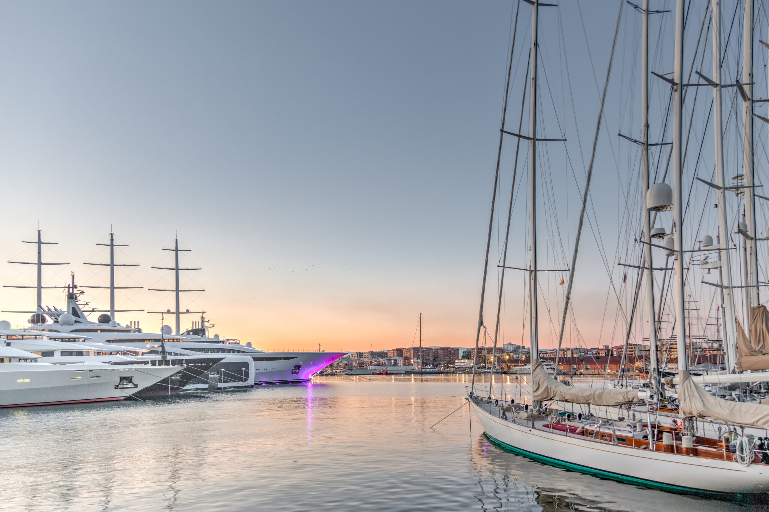 Luxury yachts and sailboats docked in a marina at sunset with city buildings in the background.