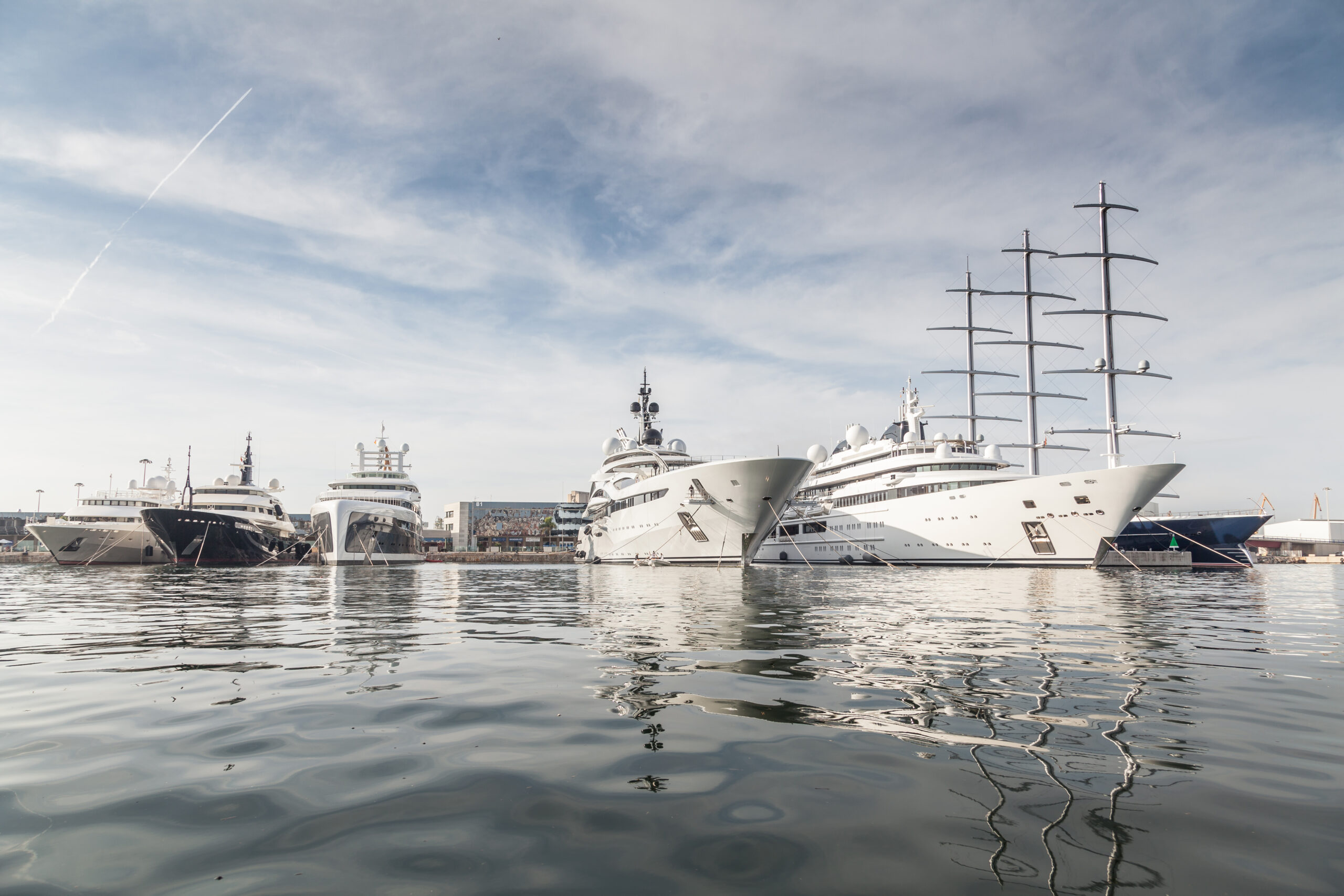 Several large luxury yachts are docked side by side in a calm marina under a partly cloudy sky.