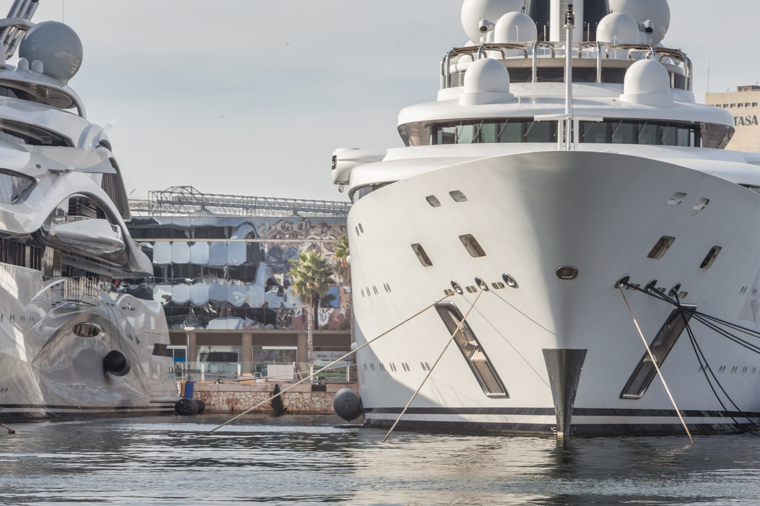 Two large luxury yachts are docked side by side in a marina with modern buildings in the background.