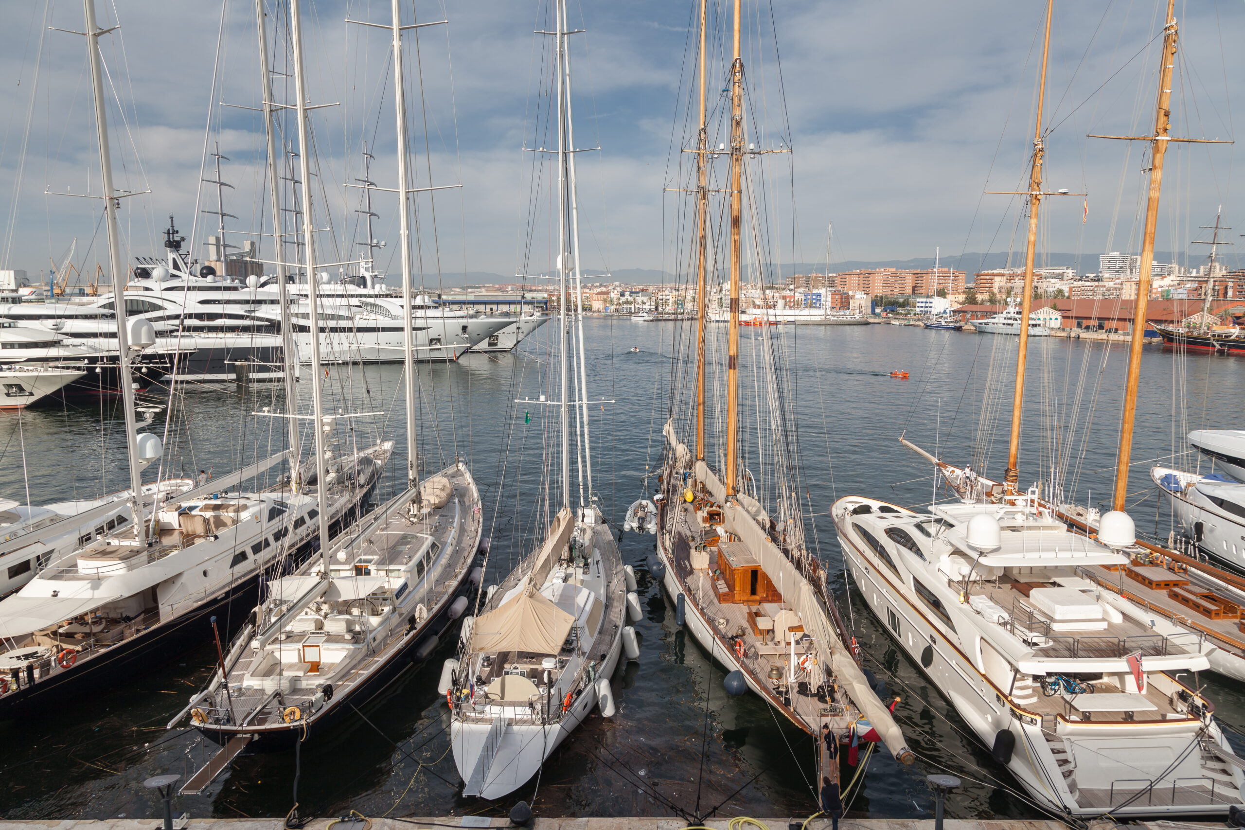 Several large sailboats and luxury yachts docked in a marina with a coastal city in the background.