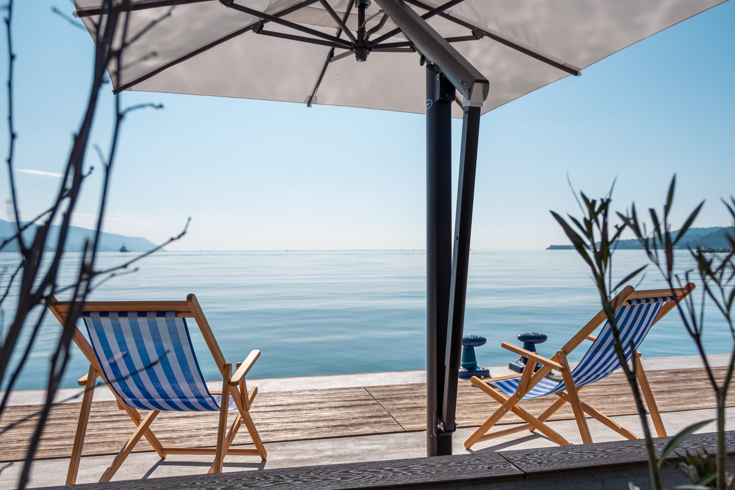 Two striped deck chairs under a large parasol on a megayacht quay overlooking a calm sea.