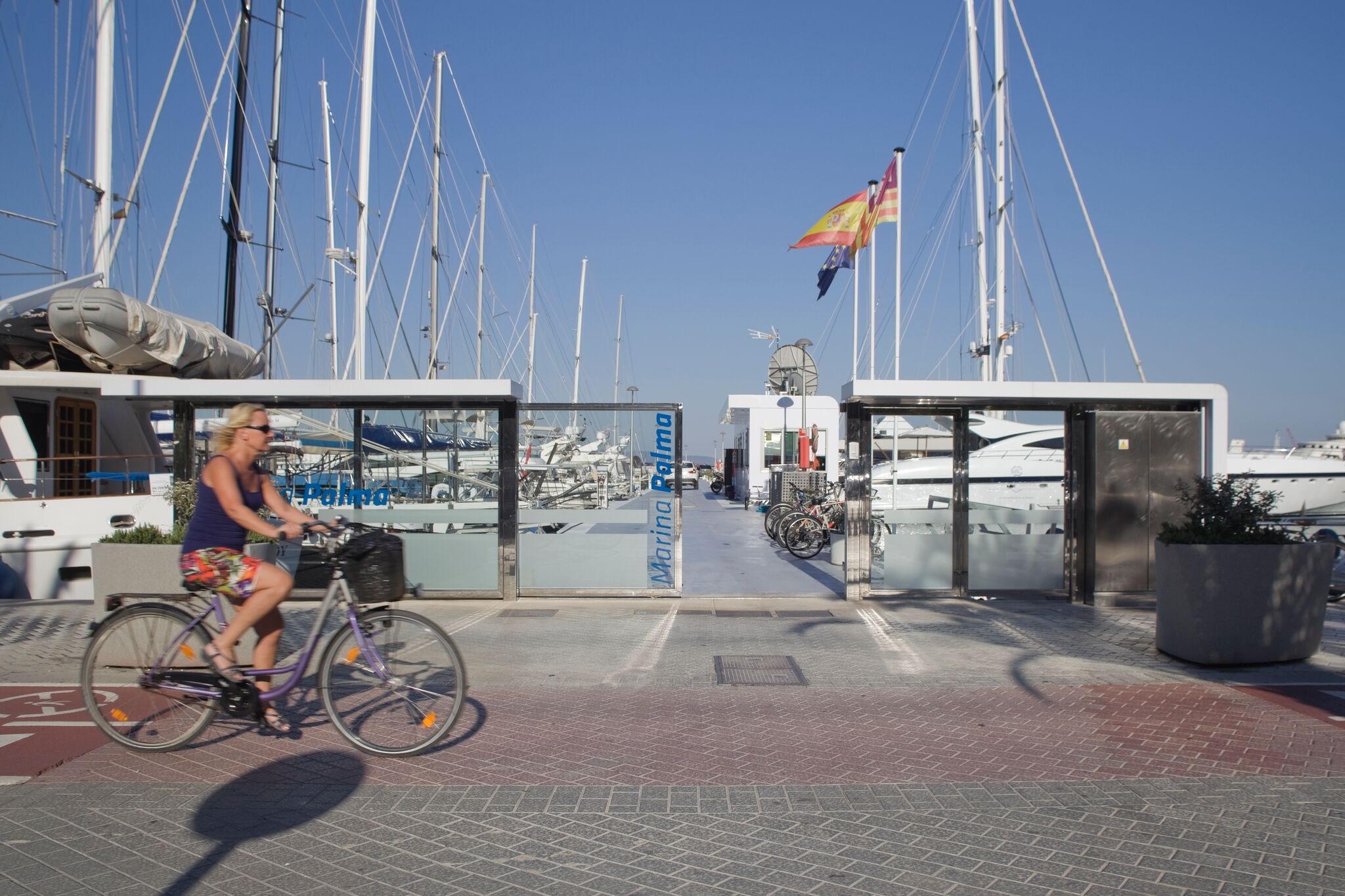 A woman rides a bicycle past the entrance gate of Marina Palma, with yachts and sailboats docked in the background.