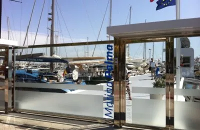 View of a marina through the glass entrance of Marina Palma, with yachts and sailboats docked under a sunny sky.