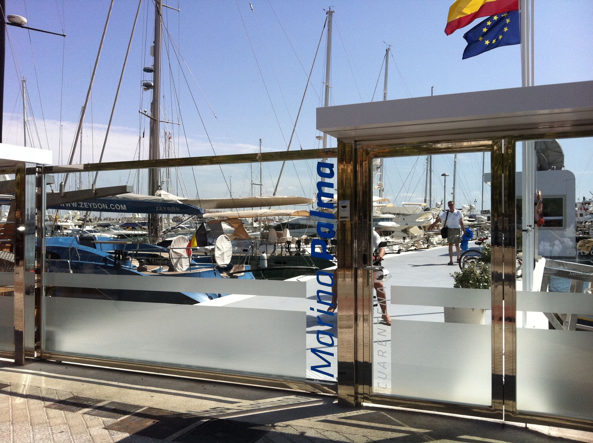 View of a marina through the glass entrance of Marina Palma, with yachts and sailboats docked under a sunny sky.