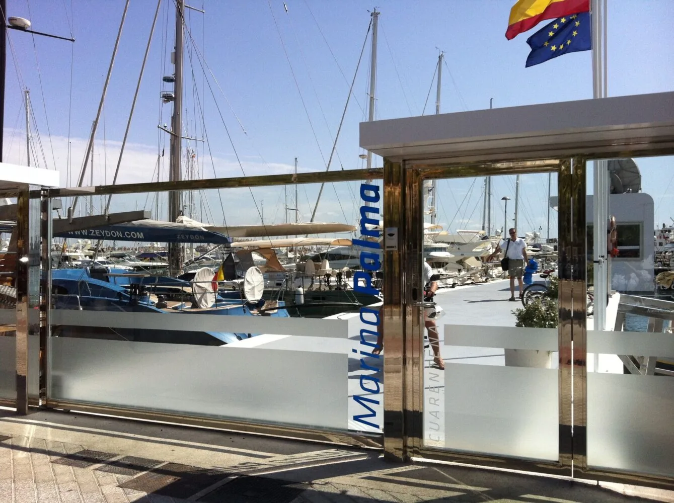 View of a marina through the glass entrance of Marina Palma, with yachts and sailboats docked under a sunny sky.