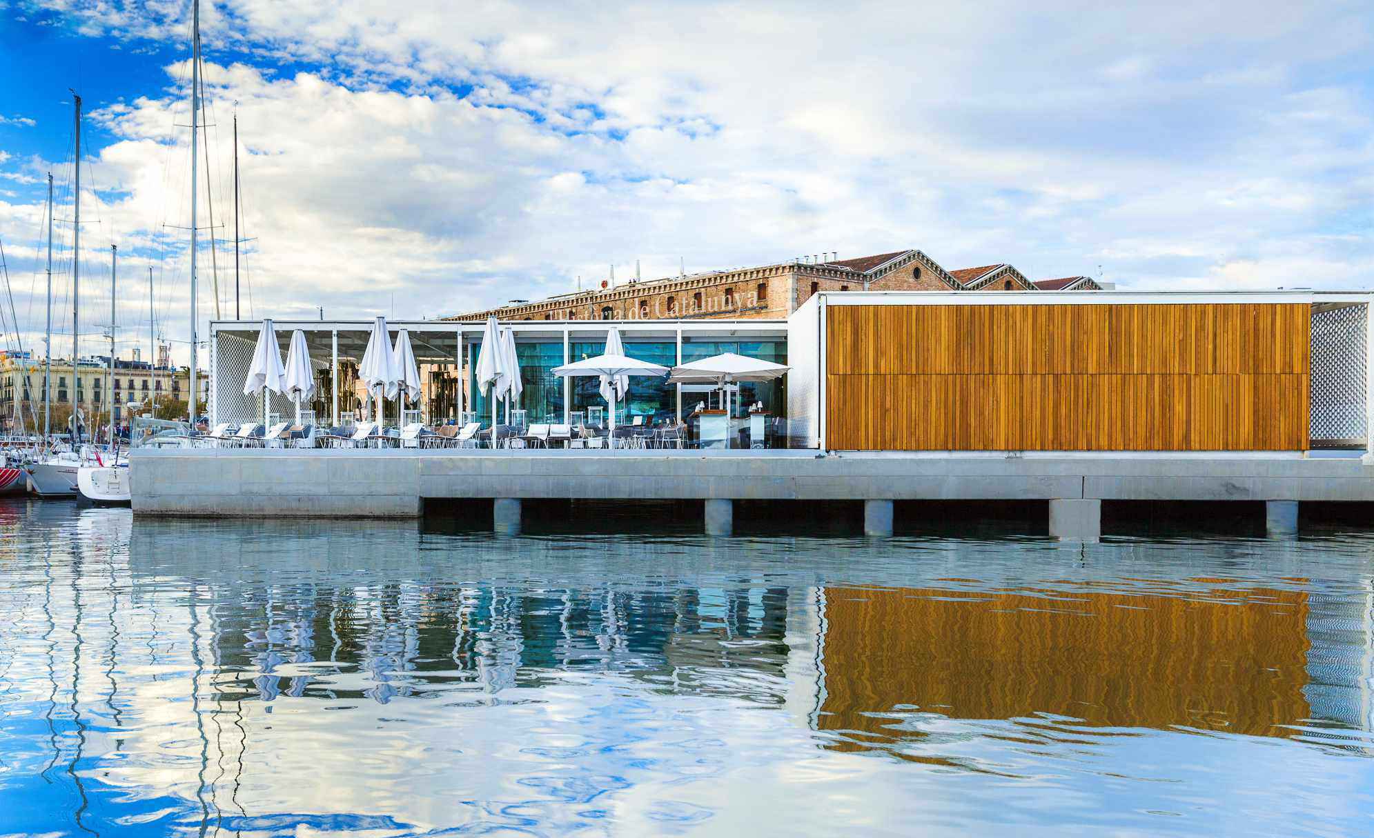 Modern waterfront building MPVR with outdoor seating and parasols reflected in the calm harbor water.