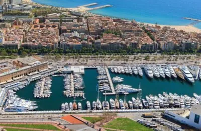 Luftaufnahme des Yachthafens Port Vell in Barcelona, auf der zahlreiche Luxusyachten zu sehen sind, die entlang des Hafens vor Anker liegen, während im Hintergrund die Stadt und der Strand zu erkennen sind.