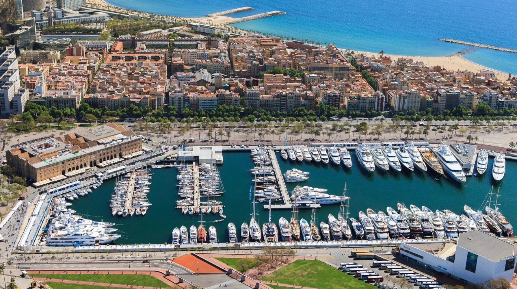 Luftaufnahme des Yachthafens Port Vell in Barcelona, auf der zahlreiche Luxusyachten zu sehen sind, die entlang des Hafens vor Anker liegen, während im Hintergrund die Stadt und der Strand zu erkennen sind.