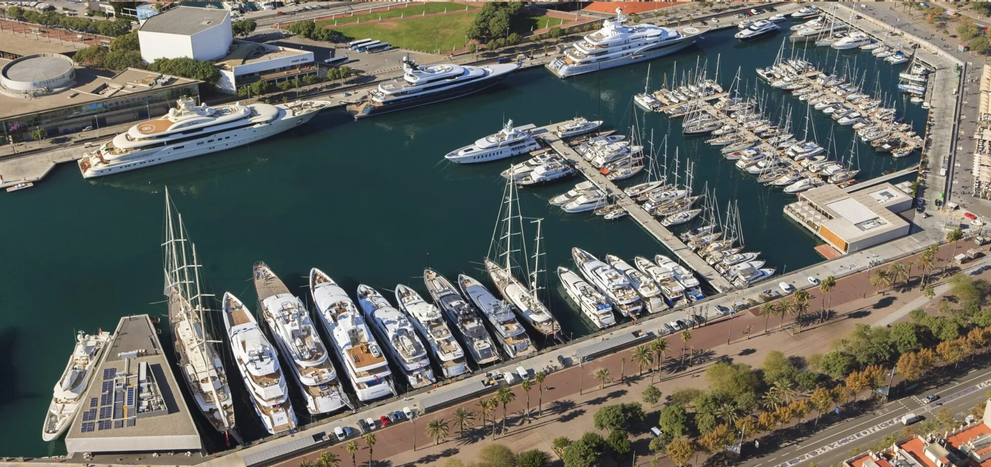 Aerial view of Marina Port Vell in Barcelona, filled with luxury yachts and surrounded by waterfront buildings and palm-lined streets.
