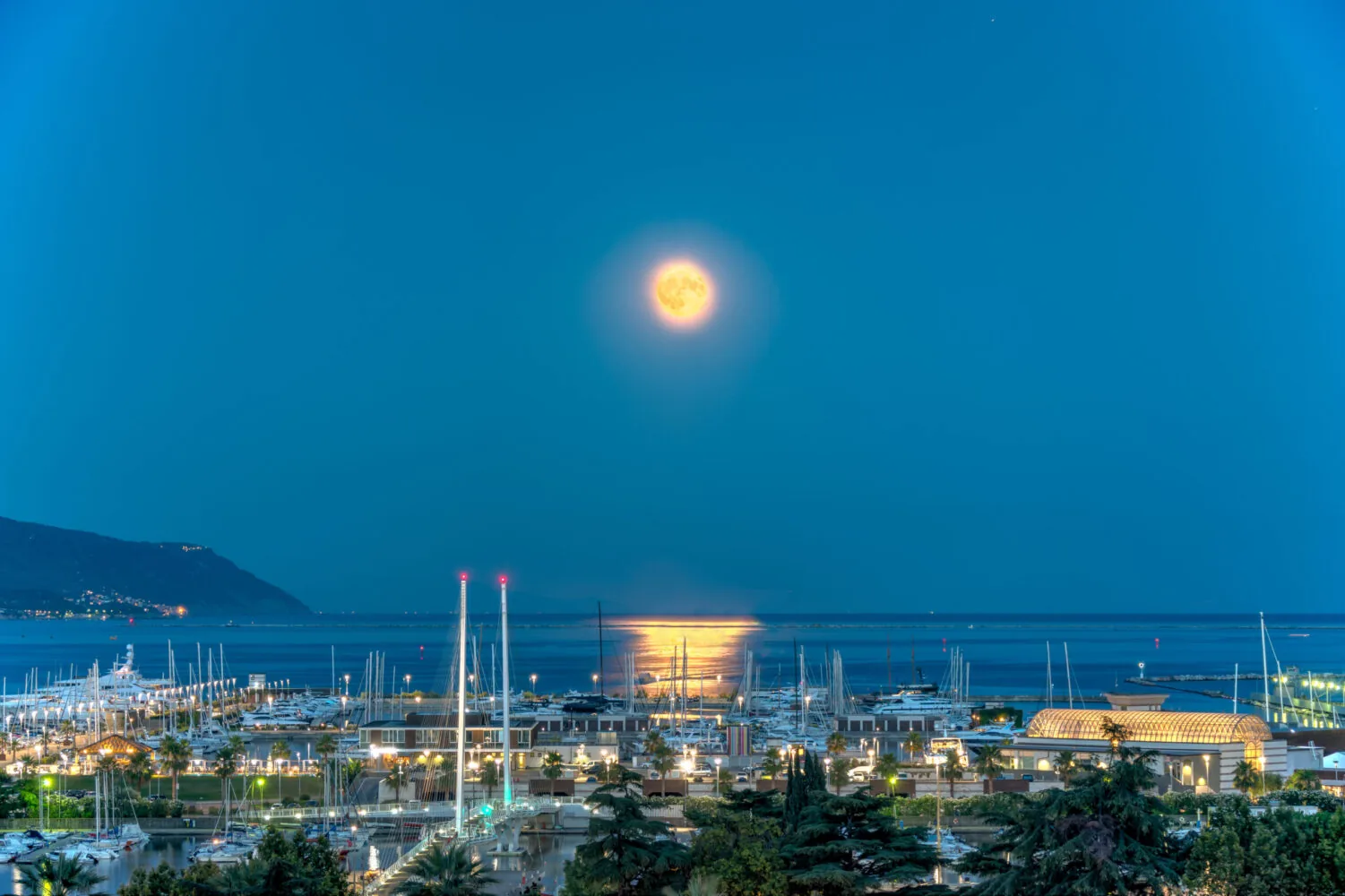 Panoramic view of a marina at night with a full moon reflecting on the sea, surrounded by boats and a lit-up waterfront.