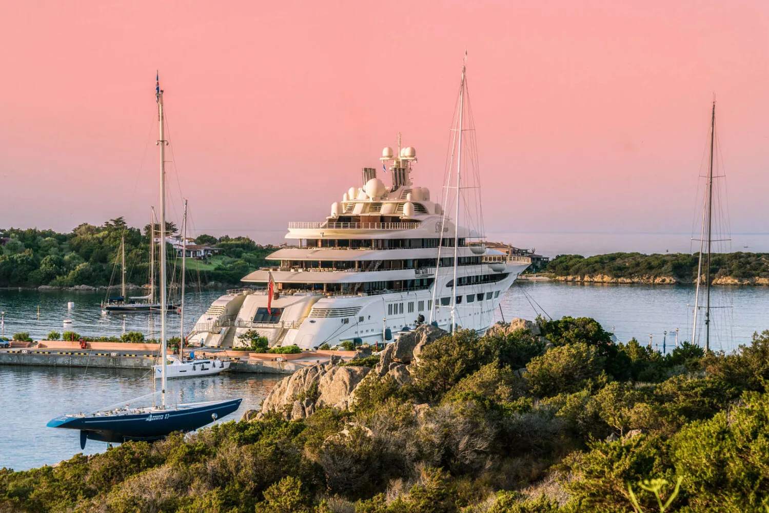 Luxe jacht aangemeerd in Porto Cervo Marina met zeilboten en weelderig groen onder een roze zonsondergang.