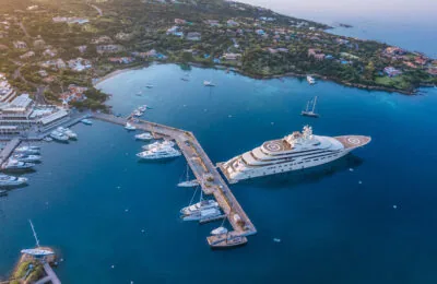 Aerial view of Porto Cervo Marina with luxury yachts docked along the pier in a scenic coastal setting.