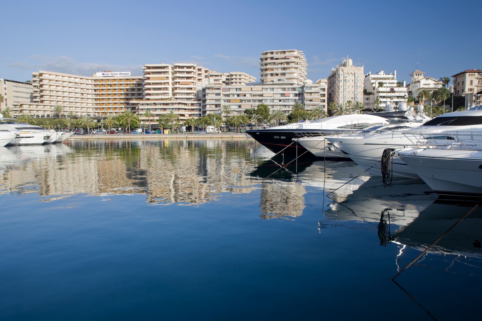 Luxury yachts docked in a calm marina with the Melia Palas Atenea hotel and surrounding buildings in the background.